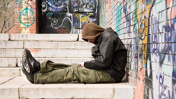 hooded homeless guy sitting on pavement against wall with grafitti